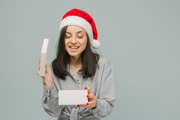 Foto der netten lächelnden Frau im Weihnachtshut öffnet Geschenk. Trägt graues Hemd, isolierten grauen Farbhintergrund.