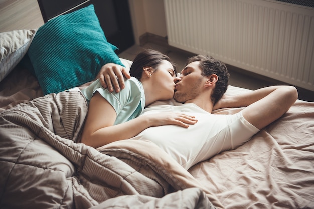 Foto der oberen Ansicht eines jungen Paares, das im Bett liegt und küsst, während es unter einer Steppdecke umarmt