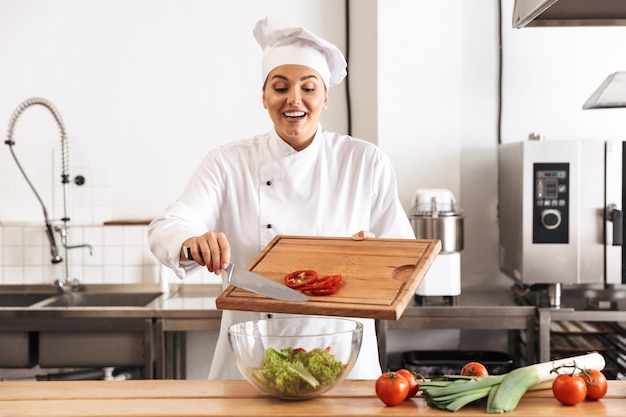 Foto der positiven Köchin, die weiße Uniform trägt, die Salat mit frischem Gemüse macht, in der Küche am Restaurant