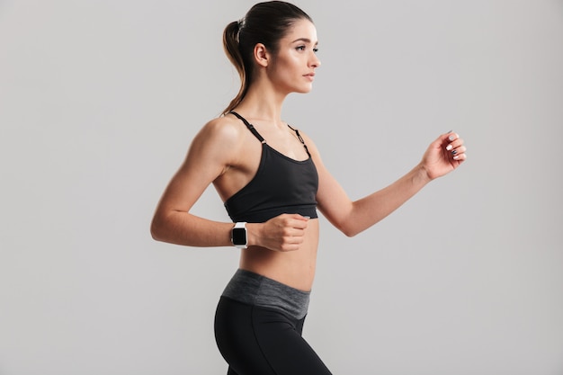 Foto der schlanken Fitnessfrau, die mit Uhr am Handgelenk läuft oder herausarbeitet, lokalisiert über graue Wand