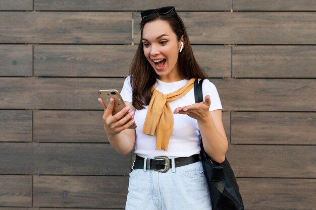 Foto der schönen erstaunlichen verblüfften jungen Frau, die Freizeitkleidung trägt, die in der Straße steht, die Kommunikation über das Mobiltelefon betrachtet Smartphone und Spaß und gute Laune habend.