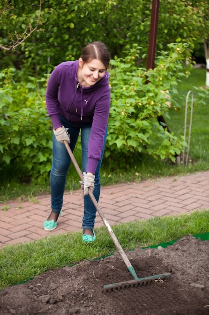 Foto der schönen Frau, die mit Rechen auf Gartenbett arbeitet