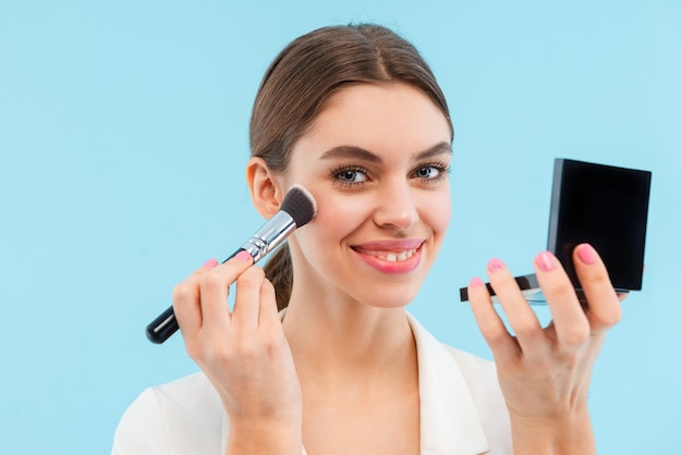 Foto der schönen jungen Frau, die isoliertes Halten Make-up Pinsel und Spiegel aufwirft.