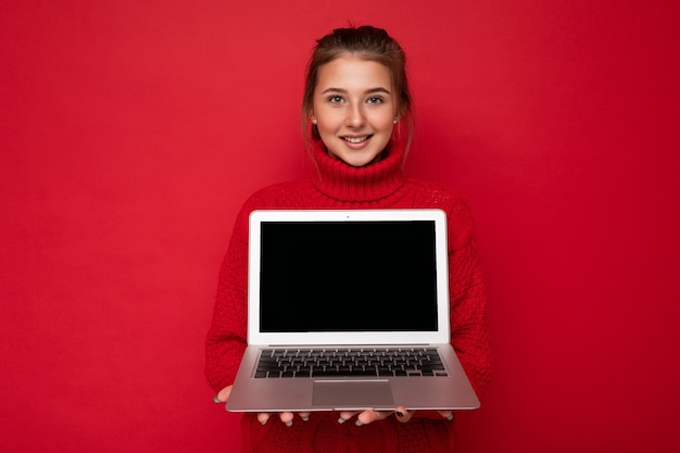 Foto der schönen zufriedenen glücklichen jungen Frau, die Computer-Laptop hält, der roten Pullover lokalisiert über roter Wand trägt. ausgeschnitten