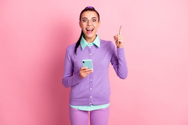 Foto der süßen Frau, die das Telefon direkt mit dem Finger nach oben offenem Mund hält, tragen Spezifikationen violette Strickjacke isoliert rosafarbener Hintergrund