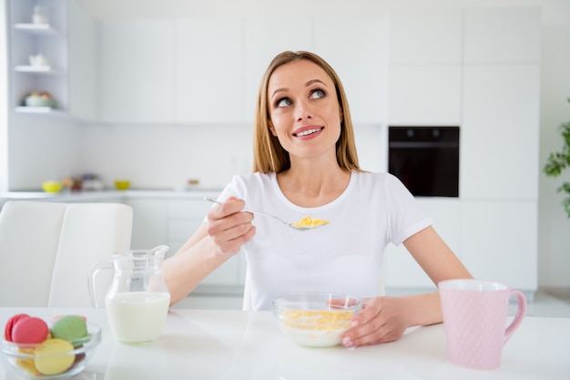 Foto der Träumerhausfrau, die Löffel hält, der Frühstück Cornflakes gesunde Ernährung Diät hält träumerisch sich vorstellt sich Kinn sitzenden Tisch weißes Licht Küche drinnen