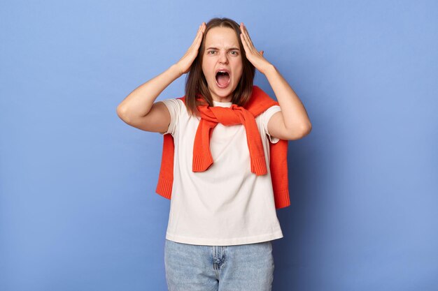 Foto der Verzweiflung schockierte wütende junge erwachsene Frau, gekleidet in ein weißes T-Shirt und einen orangefarbenen Pullover, der über die Schultern gebunden ist und vor einer blauen Wand steht und vor Hass und Wut schreit, hält den Mund offen