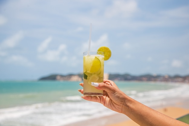 Foto der weiblichen Hand, die ein Getränk mit dem Ozean im Hintergrund hält. Urlaubskonzept