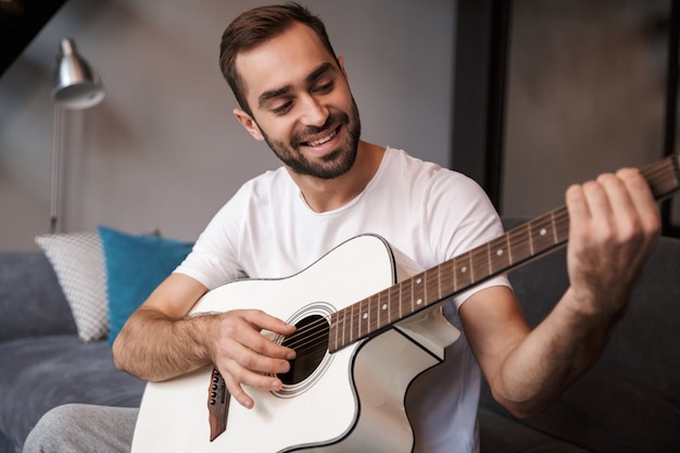 Foto des attraktiven Mannes 30s, der lässiges T-Shirt trägt, das akustische Gitarre spielt, während auf Sofa in Wohnung sitzt