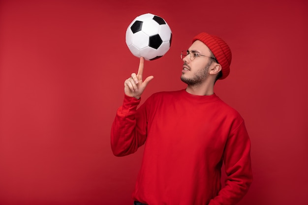 Foto des attraktiven Mannes mit Bart in den Gläsern und in der roten Kleidung. Glücklicher Mann hält den Ball auf dem Zeigefinger, isoliert über rotem Hintergrund.