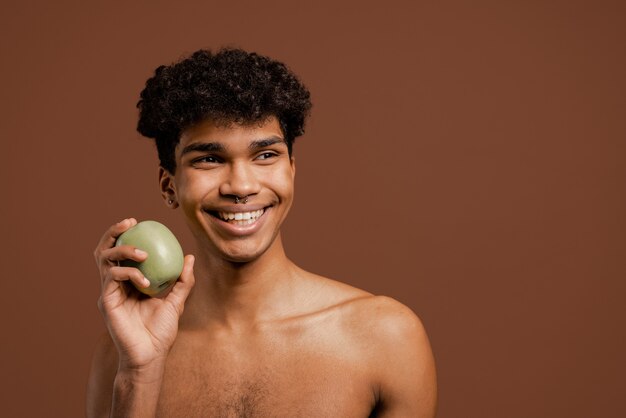 Foto des attraktiven schwarzen Mannes mit Piercing hält Apfel und lächelt. Nackter Torso, isolierter brauner Farbhintergrund.
