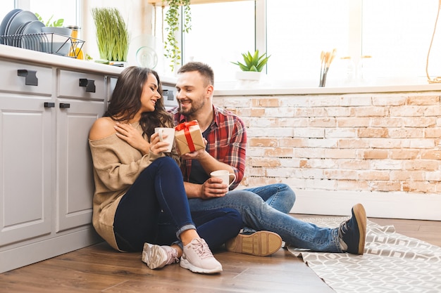 Foto des aufgeregten brünetten Paares Mann und Frau 20s beim Frühstück in der Wohnung beim Sitzen auf dem Boden mit Geschenkbox. Urlaub feiern.