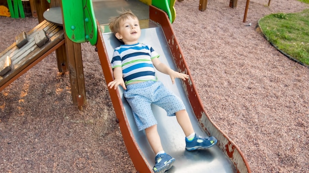 Foto des entzückenden lächelnden Jungen, der auf Folie klettert und reitet. Aktives Kind, das Spaß hat und im Park spielt