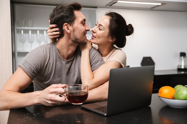 Foto des erstaunlichen Paares Mann und Frau, die in der Küche sitzen und Laptop verwenden, während sie zusammen frühstücken