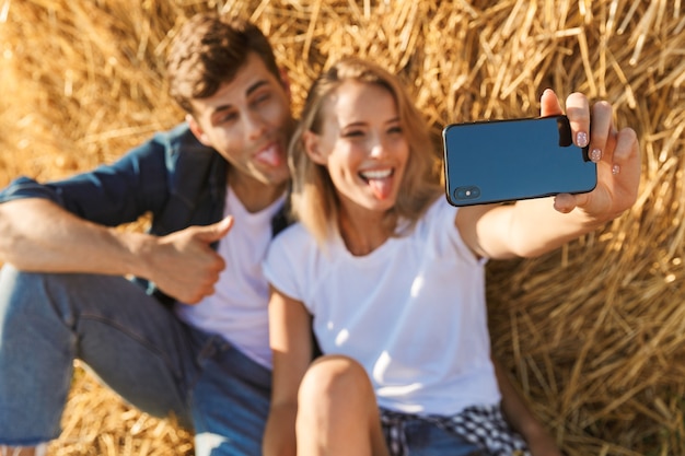 Foto des erstaunlichen Paares Mann und Frau, die selfie nehmen, während unter großem Heuhaufen im goldenen Feld, während des sonnigen Tages sitzen