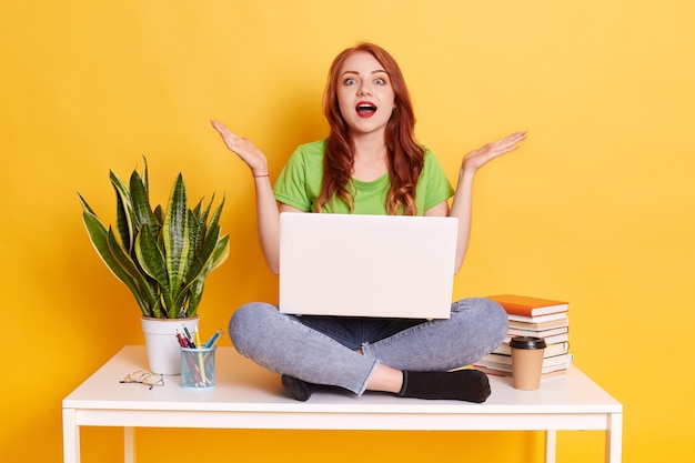 Foto des erstaunten Mädchens sitzen auf Tisch mit gekreuzten Beinen, arbeitet über Laptop, trägt grünes Hemd und Jeans, posiert isoliert auf gelber Wand, spreizt Hände beiseite, rothaariges Mädchen mit geöffnetem Mund.