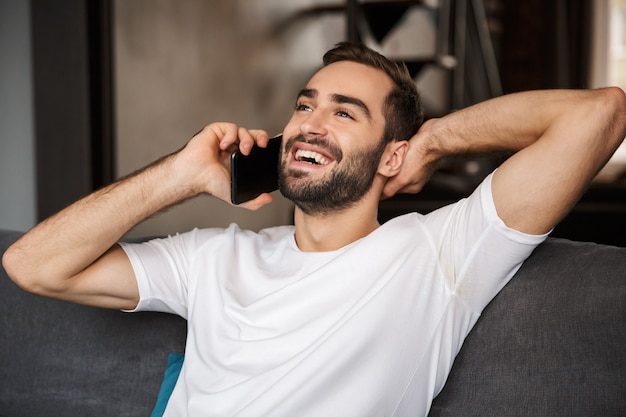 Foto des freudigen Mannes 30s, der lässiges T-Shirt trägt, das auf Smartphone spricht, während auf Sofa im Wohnzimmer sitzt
