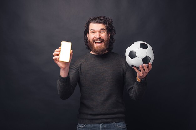 Foto des fröhlichen jungen gutaussehenden bärtigen Mannes, der Fußball hält und leeren Bildschirm auf Smartphone zeigt