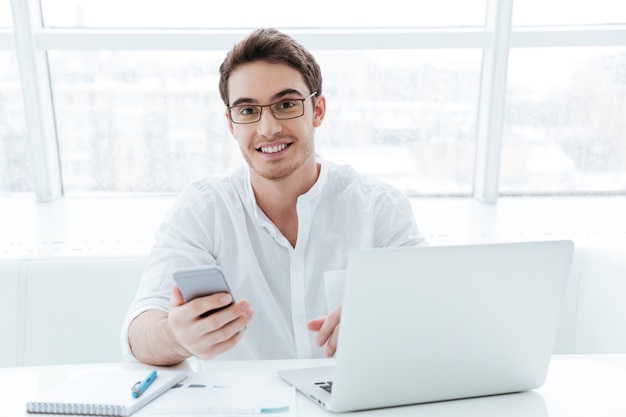 Foto des fröhlichen jungen Mannes im weißen Hemd mit Laptop-Computer beim Chatten gekleidet. Schauen Sie in die Kamera.