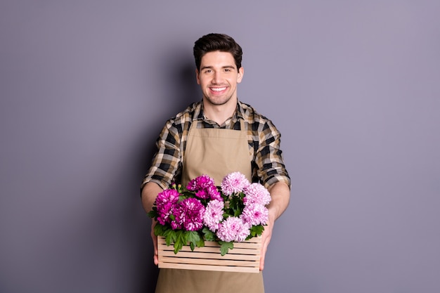 Foto des fröhlichen positiven Mannes, der zahnig arbeitet, der als Florist arbeitet, der Blumen liefert, befahl, sie zu yous isolierter grauer Farbwand zu geben