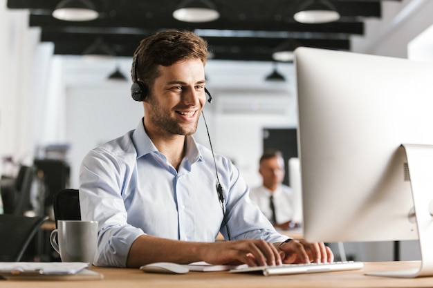 Foto des geschäftsmäßigen Mannes 20s, der Bürokleidung und Headset trägt und am Computer im Callcenter arbeitet