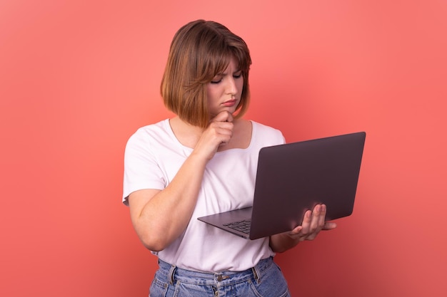 Foto des gesinnten freiberuflichen Mädchens hält Laptop-Fingerkinnabnutzungs-weißes T-Shirt rosa Farbhintergrund