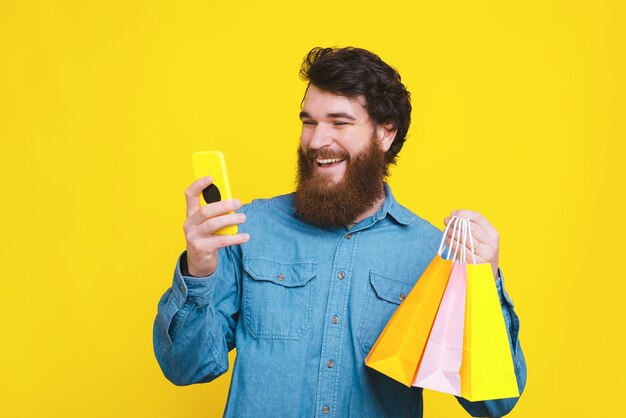 Foto des glücklichen bärtigen Hipster-Mannes, der Smartphone verwendet und Einkaufstaschen hält