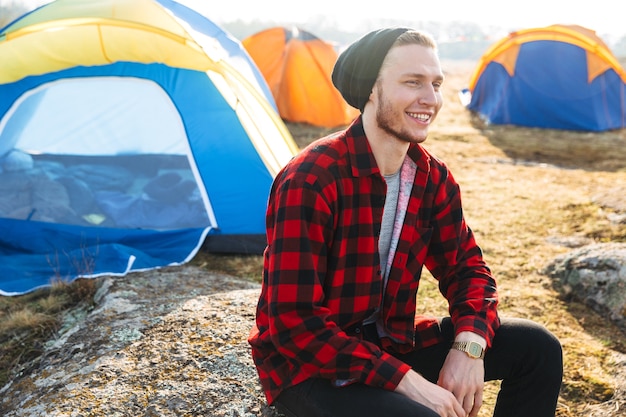 Foto des glücklichen jungen Mannes draußen mit Zelt im freien alternativen Feriencamping über den Bergen.