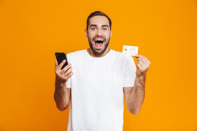 Foto des glücklichen Mannes 30s in der Freizeitkleidung, die Smartphone und Kreditkarte hält, lokalisiert