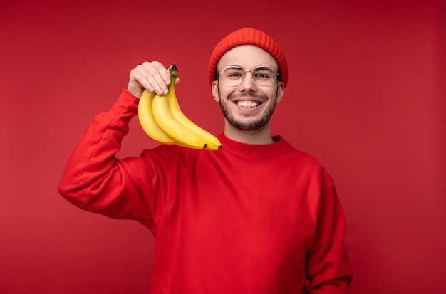Foto des glücklichen Mannes mit Bart in den Gläsern und in der roten Kleidung. Hält Bananen und lächelt, gesunder Lebensstil, isoliert über rotem Hintergrund.