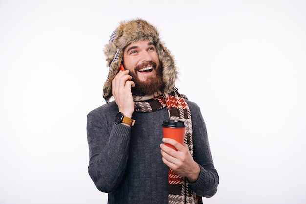 Foto des glücklichen Mannes mit Bart in der Winterkleidung, die am Telefon spricht und Tasse Kaffee hält, um zu gehen