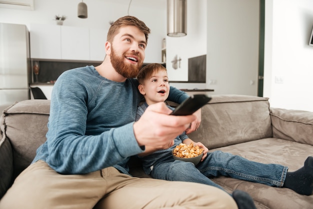 Foto des glücklichen Vaters mit Fernbedienung beim Fernsehen mit seinem kleinen süßen Sohn, der Popcorn hält.