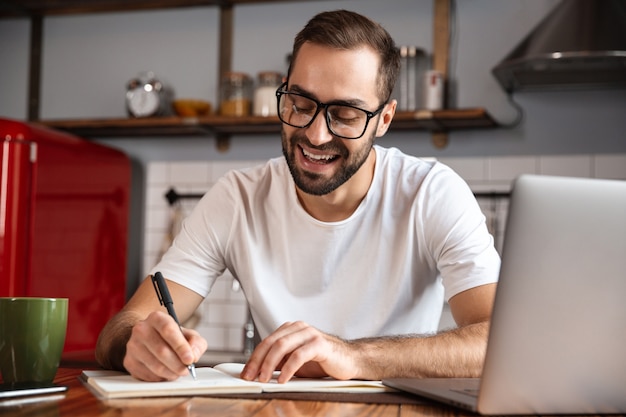 Foto des gutaussehenden Mannes 30s, der Brillen trägt, die Notizen aufschreiben, während sie silbernen Laptop auf Küchentisch verwenden
