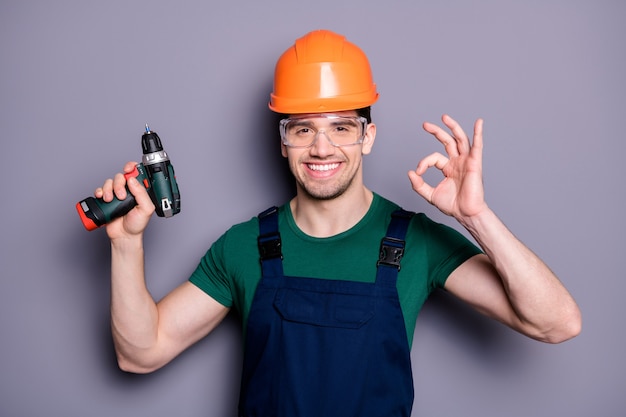 Foto des hübschen männlichen Arbeiters Kerl qualifizierter Ingenieur halten elektrische Bohrmaschine, die okey Symbol tragen T-Shirt Sicherheit Latzhose Schutzhelm Brille isoliert graue Wand zeigt