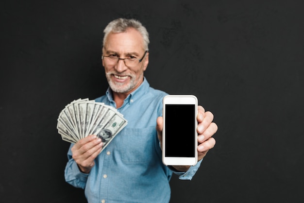 Foto des Inhalts pensionierter Mann 60s mit grauem Haar, das Fan des Gelddollar-Bargeldes hält und Handy demonstriert, lokalisiert über schwarzer Wand
