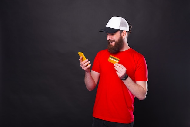 Foto des jungen freudigen bärtigen Hipster-Mannes im roten T-Shirt, der etwas online mit Kreditkarte und Smartphone kauft