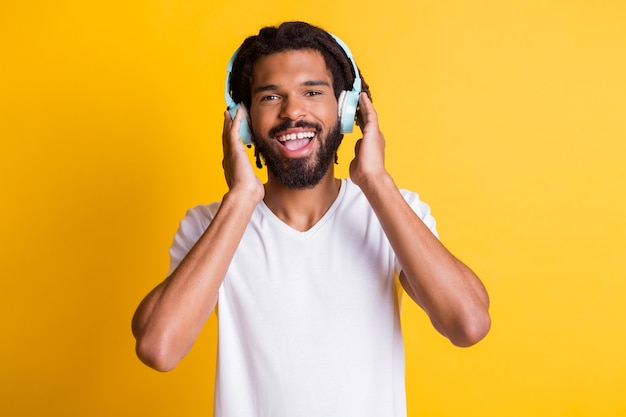 Foto des jungen schwarzen Kerls, der Kopfhörer mit offenem Mund und einem zahnigen Lächeln hält, trägt Kopfhörer weißes T-Shirt isoliert gelber Farbhintergrund
