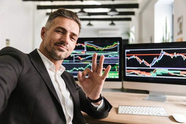 Foto des kaukasischen mannes 30s, der anzug trägt, der selfie nimmt, während im büro am computer mit grafiken und diagrammen am bildschirm arbeitet