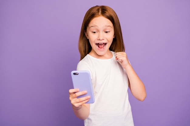 Foto des lustigen kleinen aufgeregten Ingwermädchens, das Telefon erhöht Faust erhöht