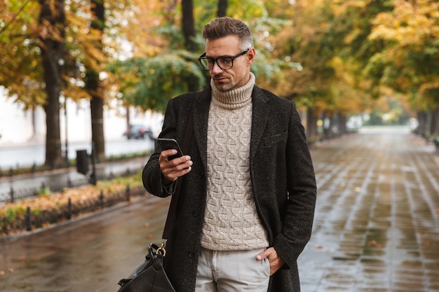 Foto des Mode-Macho-Mannes der 30er Jahre, der warme Kleidung trägt, die draußen durch Herbstpark geht und Handy benutzt