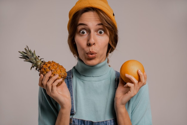 Foto foto des niedlichen weiblichen hipsters hält ananas und orange mit überraschung, sieht gesund aus. weiße frau trägt jeans insgesamt und hut isolierten grauen farbhintergrund.