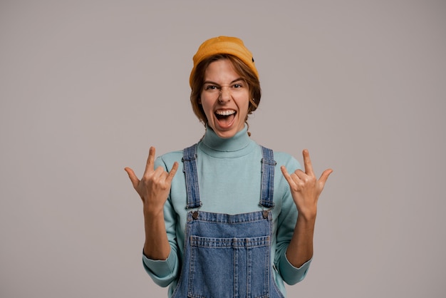 Foto des niedlichen weiblichen Hipsters zeigt Geste Rock'n'Roll. Weiße Frau trägt Jeans insgesamt und Hut isolierten grauen Farbhintergrund.