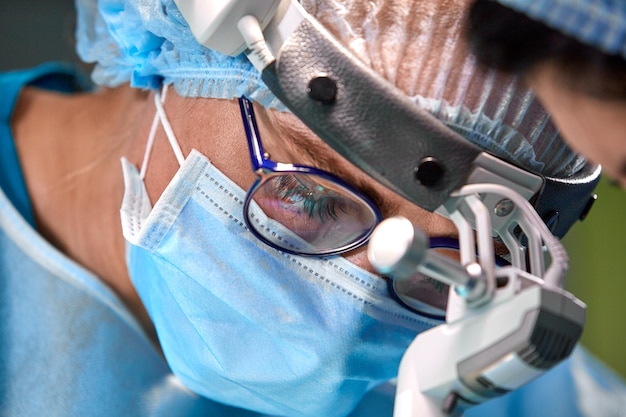 Foto des operierenden Chirurgen im OP-Saal Chirurg mit Maske und Brille mit montiertem Scheinwerfer Porträt schließen