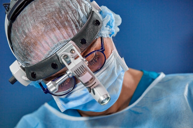 Foto des operierenden Chirurgen im OP-Saal Chirurg mit Maske und Brille mit montiertem Scheinwerfer Porträt schließen