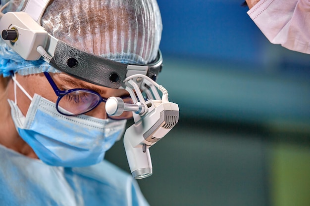 Foto des operierenden Chirurgen im OP-Saal Chirurg mit Maske und Brille mit montiertem Scheinwerfer Porträt schließen