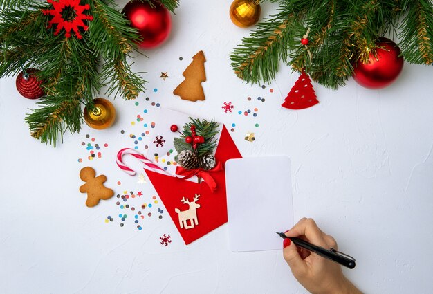 Foto des roten Weihnachtsumschlags, der Kekse, der Hände mit Stift, der Zweige der Weihnachtsdekoration der Fichte auf leerem Weiß