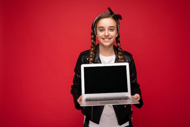 Foto des schönen jungen Mädchens, das den Computerlaptop hält, der die Kamera lokalisiert über buntem Hintergrund betrachtet.