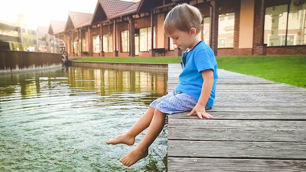 Foto des süßen 3-jährigen kleinen Jungen, der am Ufer des Flusses am Wasserkanal in der Altstadt sitzt und mit den Füßen Wasser spritzt.