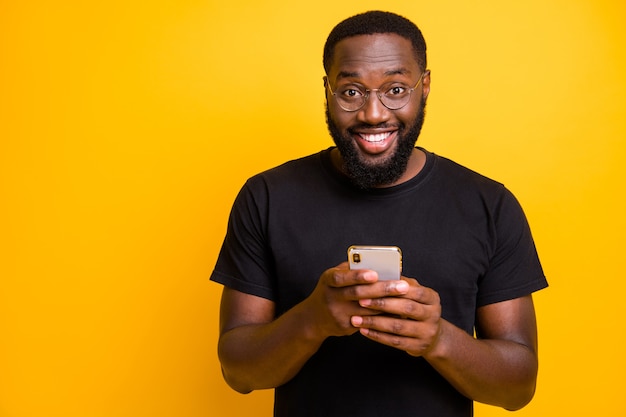 Foto des trendigen fröhlichen positiven gutaussehenden Mannes, der zahnlos in Brillen lächelt, die durch sein Telefon in der isolierten lebendigen Farbwand des T-Shirts durchsuchen