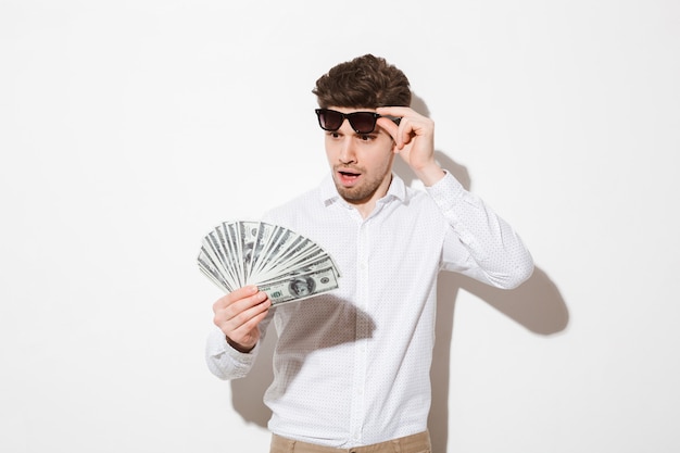 Foto des überraschten glücklichen Mannes im Hemd, das schwarze Sonnenbrille abnimmt und Fan von Gelddollar-Banknoten mit Aufregung betrachtet, lokalisiert über weißer Wand mit Schatten